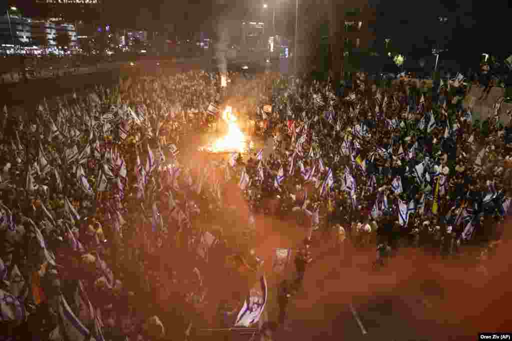 Protestuesit me flamuj në duar shihen përreth një zjarri në Tel Aviv. 